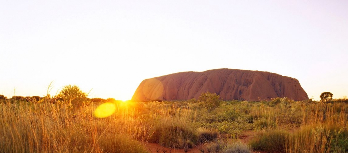 a sunset over a dry grass field with Uluru in the background